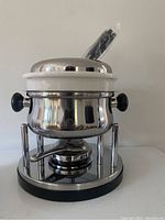 New Alpina Switzerland fondue pot with stainless steel outer body and white ceramic insert, black handles on sides, and lid with fondue forks resting inside.