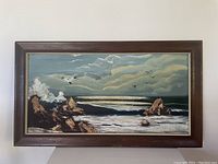 Front view of rectangular oil painting on board showing coastal landscape with rocky shore, waves, cloudy sky, and flying birds. Painting is in a wide wooden frame with dark stain and gold inner edge, frame shows some damage and needs repair.