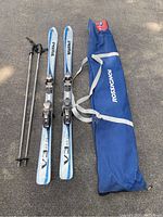 Photo shows Volkl V3 skis with bindings, ski poles, and blue Rossignol branded carry bag on pavement, with some dust visible.