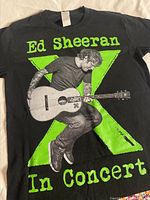Front of black Ed Sheeran tour T-shirt with image of Ed Sheeran playing guitar and 'In Concert' text.