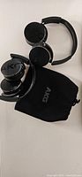 Two black AKG branded wireless over-ear headphones and one black AKG drawstring carrying pouch laid flat on a white surface.
