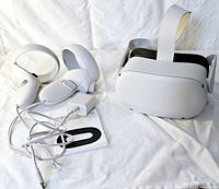 Photo showing Meta Quest 2 white VR headset with adjustable head strap, matching white wireless hand controllers, USB cable and manual laid out on white fabric background.