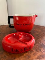 Photo showing a red water jug with black handle and Haig & Haig Scotch Whisky printed on the side, alongside a matching red ashtray with the branding visible.