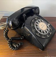 Side angle showing the black rotary dial telephone including handset, coiled cord, and base with rotary dial and number placard.