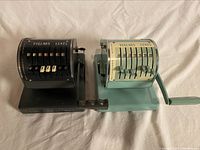 Two vintage Paymaster cheque writers side by side, black Model X-550 and blue-green Model X-900