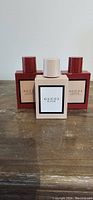 Three bottles of Gucci Bloom perfume, two Gucci Bloom Ambrosia De Fiori deep red bottles and one white Gucci Bloom bottle, arranged on a wooden surface.