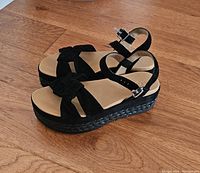 Pair of black velvet Ugg sandals with platform heel pictured on wooden floor from side angle showing buckle and straps.