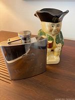Photo of polished stainless steel Wisers whiskey flask with screw cap and Staffordshire Toby Jug figurine in the background on wooden slats.