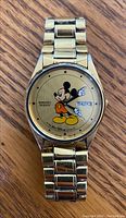 Front view showing gold-tone dial with full-color Mickey Mouse graphic, date and day window, and gold-tone link bracelet.