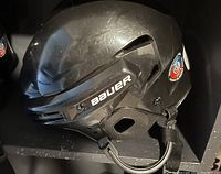 Side view of one black children's Bauer hockey helmet showing white Bauer branding, ventilation slots, adjustable chin strap, and hockey certification sticker. Helmet has surface scratch marks.