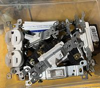 Clear plastic container holding assorted white electrical receptacles and white toggle light switches with metal mounting brackets and screws attached.