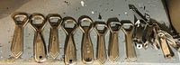 A collection of 14 assorted vintage bottle openers laid out on a surface, showing 10 similar metal wall-mounted types in a row and 4 varied designs to the right side.