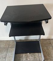 Black desk with upper working surface, keyboard tray, and lower shelf on silver metal frame, placed on tiled floor against a white wall.