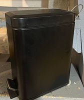 Black rectangular metal step trash can with foot pedal and hinged lid, matte black finish with minor wear marks, photographed against a stone floor and wall background.