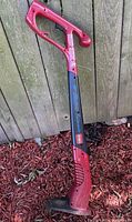 Photo of a red and black electric Toro weed trimmer leaning against a wooden fence on ground covered with mulch.