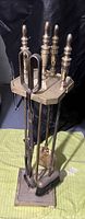 Photo shows the complete fireplace poker set including a poker, shovel, broom, tongs, all standing in a metal stand. Decorative handles visible on tools. Metal is aged with a patina.