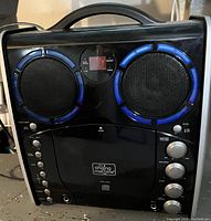 Front view of black portable karaoke machine showing built-in stereo speakers with blue accents, CD player door with The Singing Machine logo, multiple control knobs for audio adjustments, and integrated handle.