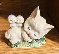 Porcelain figurine of a white kitten sleeping with two small birds on its back, resting on a base that resembles green leaves.