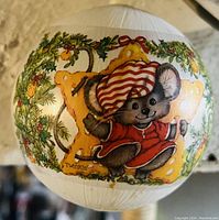 Photo of round white Christmas ornament featuring a smiling mouse named Twinkle wearing a red outfit with striped nightcap, surrounded by holly, bells, and festive greenery.