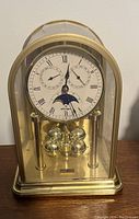 Photo of a German-made brass table top clock under glass dome, showing white dial with Roman numerals, moon phase dial, and pendulum with four brass balls.