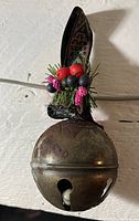 Photo of a single round brass Christmas bell with patina, decorated with faux holly leaves, berries, and a black ribbon loop on top.
