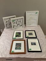 Overview of six small framed art pieces on a light pink cloth background against grey wall, featuring two butterfly-themed shadow-box frames, a gold lettering quote print, and four smaller floral and scenic prints in varying frame styles.