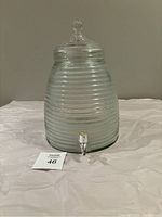 Glass beehive shaped beverage dispenser with metal spigot on a white surface against grey background.