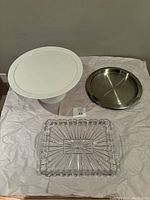 Front view of all three items together: white cake stand, round metal tray, and rectangular glass tray.