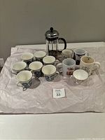 Overall shot showing all 12 coffee mugs and French press coffee maker on a cloth surface.