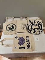 Four cotton tote bags laid out, showcasing different literary prints. Brain tote has matching pin and pop socket accessories.