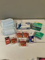 Photo showing disposable blue surgical face masks stacked, Vguard nitrile gloves box, multiple Medi Care flexible fabric bandage boxes, antiseptic towelettes, and bandage strips.