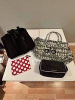 Overview of the tote bags and pouches showing the black neoprene B HalfMoon City Tote, the gray and white Alo tote bag, the red polka dot zipper pouch, and the black Victoria's Secret garment bag on a white table.