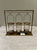 Front view of the hanging tri-candle holder showing three clear glass containers hanging from a bronze or brass colored metal frame on a flat base.