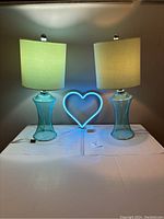 Photo showing pair of turquoise bedside table lamps lit on a white table with neon blue heart light between them.