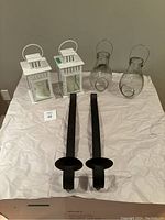 Six large candle holders displayed on white sheet. Two white metal lantern candle holders with glass sides and handles, two clear glass hurricane style holders, and two tall black metal hanging candle holders with a flat base and handle. All items visible in the photo.