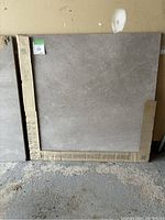 Photo of one large square grey ceramic floor tile with original cardboard partial packaging, showing size and surface texture.