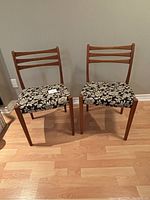 Two teak dining chairs with black floral upholstery seen from front angle, placed on light wood floor against taupe wall.
