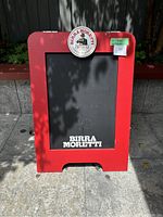Front view of red-framed metal sandwich board with black chalkboard surface and Birra Moretti logo and text