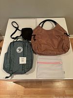 All three bags including Fjallraven Kanken backpack, Herschel crossbody, and pink IndigoWork tote laid out on table.