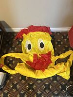 Yellow and red deflated inflatable tube man laid out on a carpeted floor, showing face and arms with cartoonish eyes and mouth.