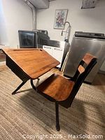 Side view of antique wooden school desk with attached chair, showing metal legs and wooden surfaces.