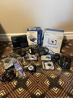 Overview of four cameras, branded boxes, chargers, cables and carrying case on patterned carpet