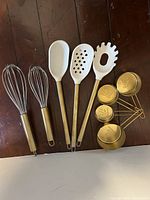 Photo showing two whisks with gold handles, three white handled spoons with gold handles, and four gold measuring cups nesting together.