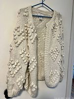 Front side view of white knit sweater with bobble heart pattern hanging on plastic hanger against wall, showing texture and fit.