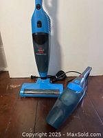 Two Bissell vacuums side by side: one upright corded vacuum and one handheld vacuum in blue and black colors shown clearly on wooden floor.