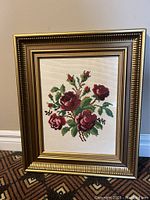 Full view of vintage framed embroidery picture with bouquet of red roses and green leaves on beige fabric background.