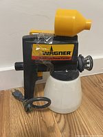 Front side of Wagner Series 100 paint sprayer showing the yellow funnel lid, black body with logo, and white paint container.