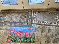 Three entrance carpets laid out on a tiled floor near glass doors; includes large beige floral patterned carpet, smaller beige patterned carpet, and brightly colored tulip print carpet.