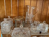 Photo showing the glass jug, butter dish, sugar and creamer set, bowl, vase, and two jars on a glass tray against a wood backdrop.