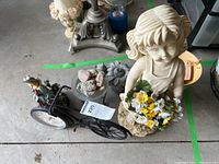 Photo showing overall lot with resin girl figurine, metal bicycle clock, and multiple garden decor pieces on a concrete floor.